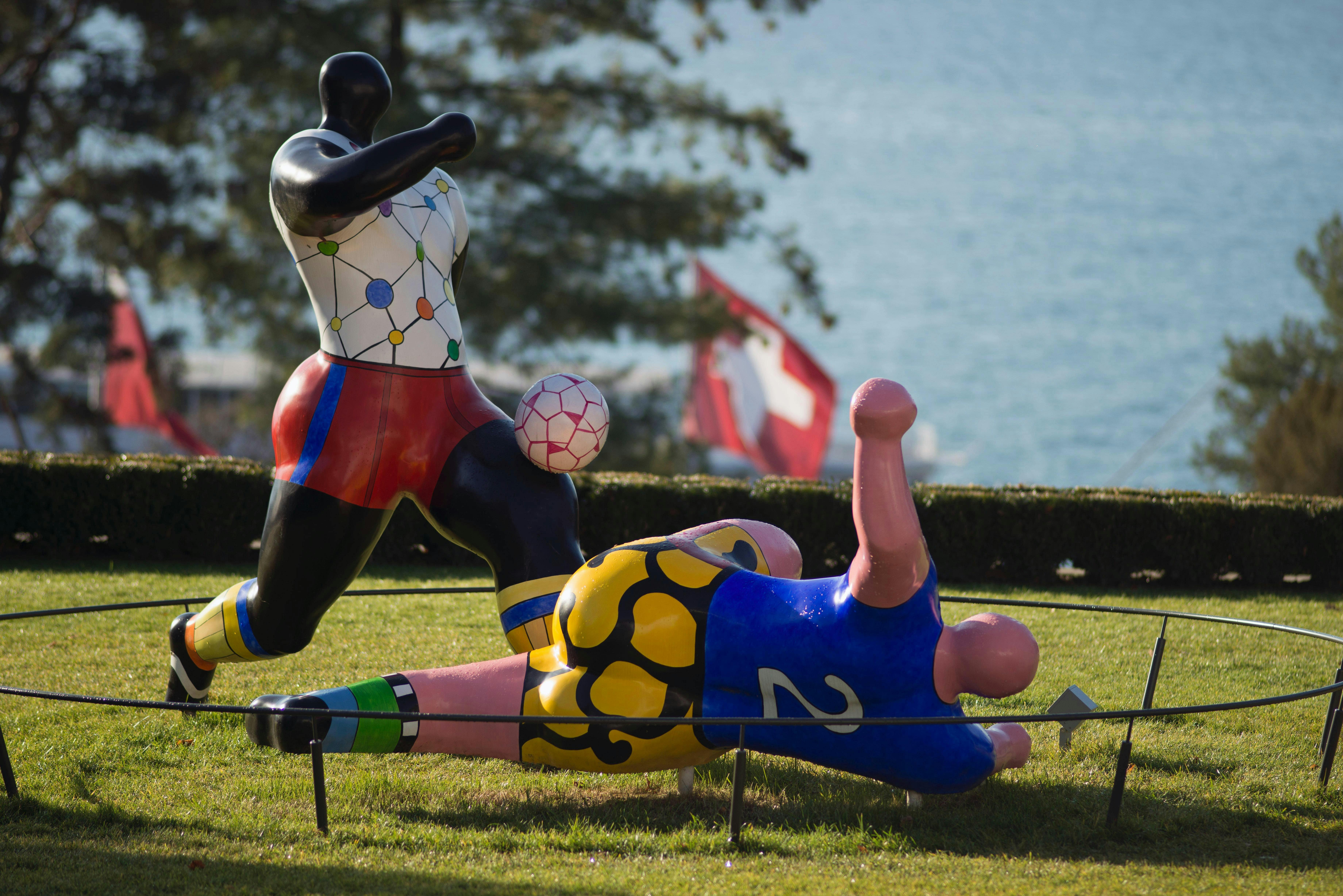 Dvě barevné, abstraktní sochy fotbalistů v akci, s jezerem a vlajkou v pozadí.