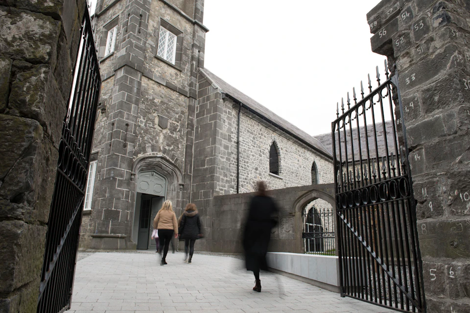 Three people walking towards an old stone church building through an open wrought iron gate.