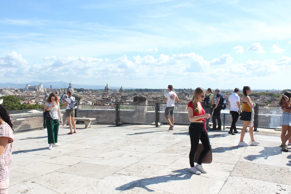 People on a large stone terrace overlooking a cityscape, with a clear sky and scattered clouds in the background.