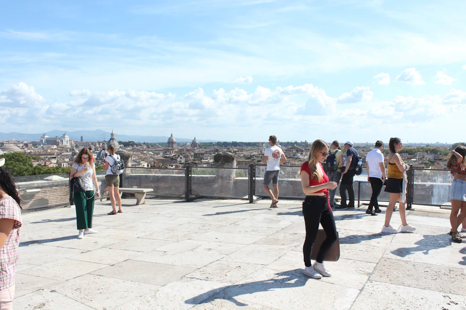 People on a large stone terrace overlooking a cityscape, with a clear sky and scattered clouds in the background.