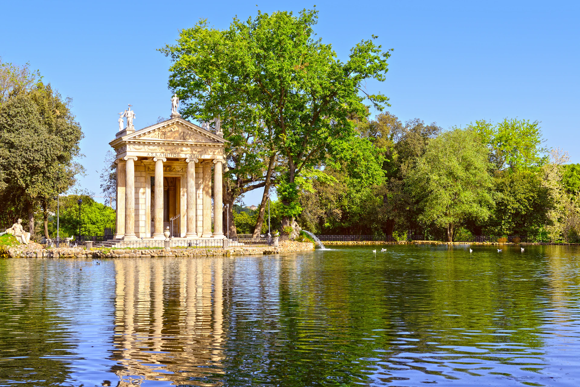 Temple-like structure near a large tree by a calm lake under a clear blue sky.