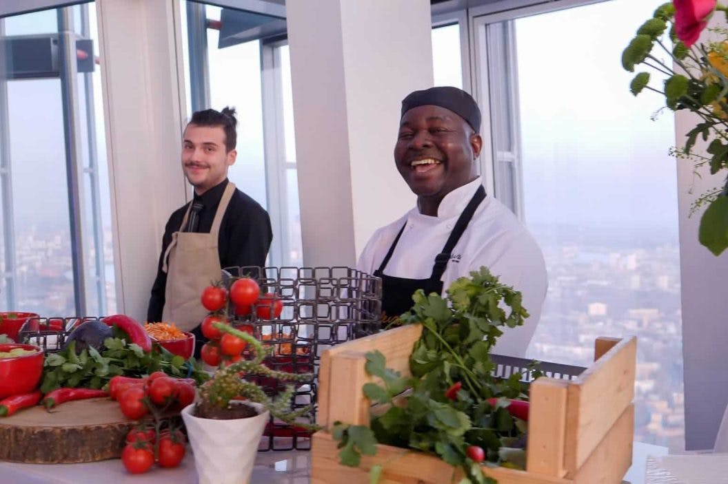 Dwóch szefów kuchni uśmiecha się do blatu kuchennego wypełnionego świeżymi warzywami i ziołami, na tle dużych okien z widokiem na miasto.