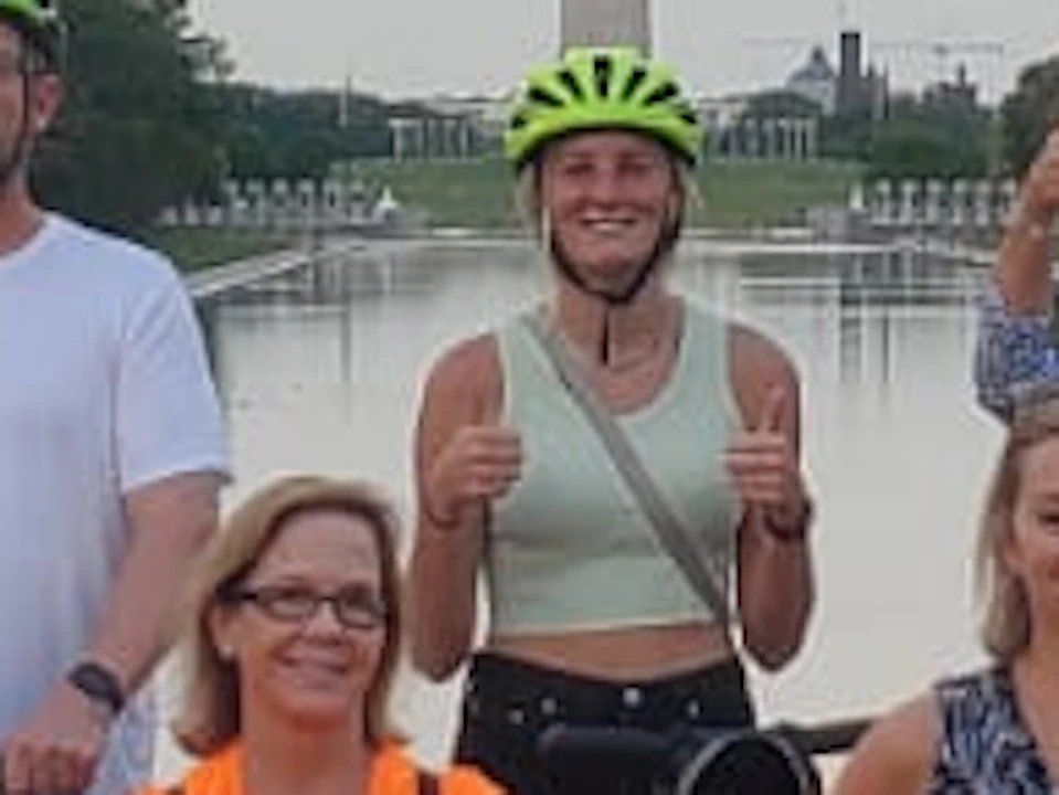 A group of people with bicycles and helmets pose in front of the Washington Monument on a cloudy day.