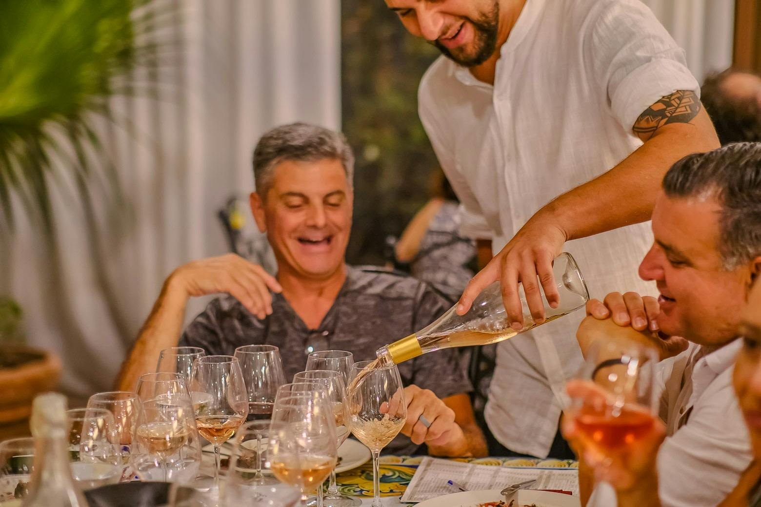 A man pours wine into glasses at a table with others seated, engaging in conversation and enjoying the moment.