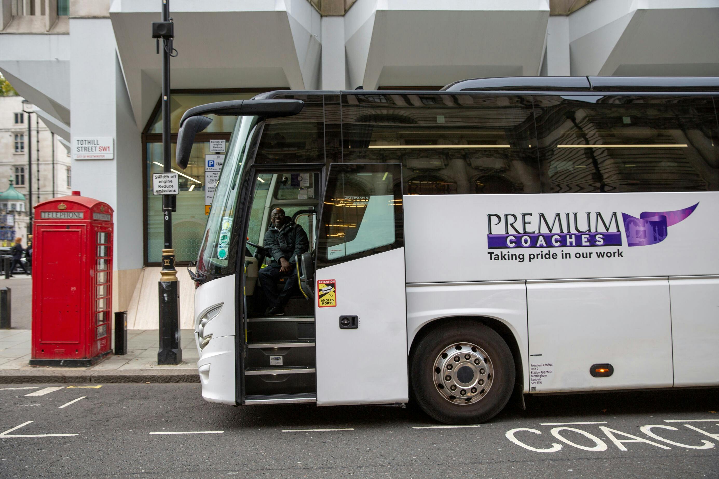 A white coach bus with its door open, parked on the side of a street. A person is visible sitting inside the driver's seat.