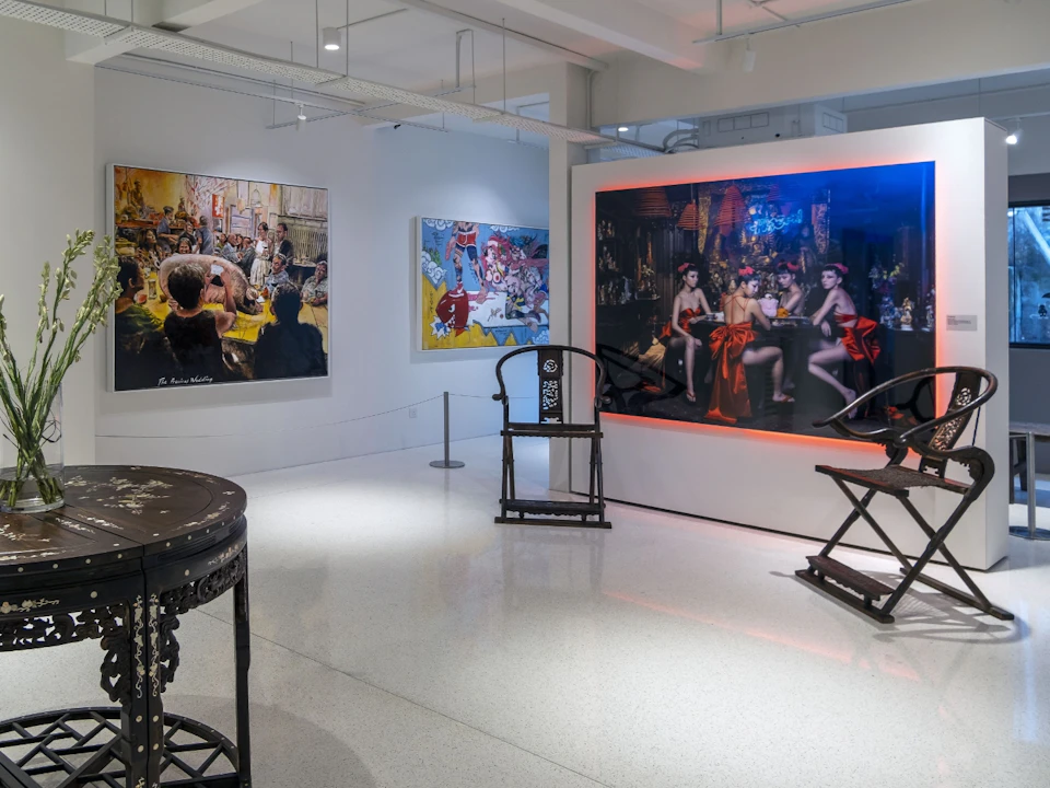 An art gallery interior with paintings on the walls, furniture pieces, and a polished floor. One artwork is brightly lit.