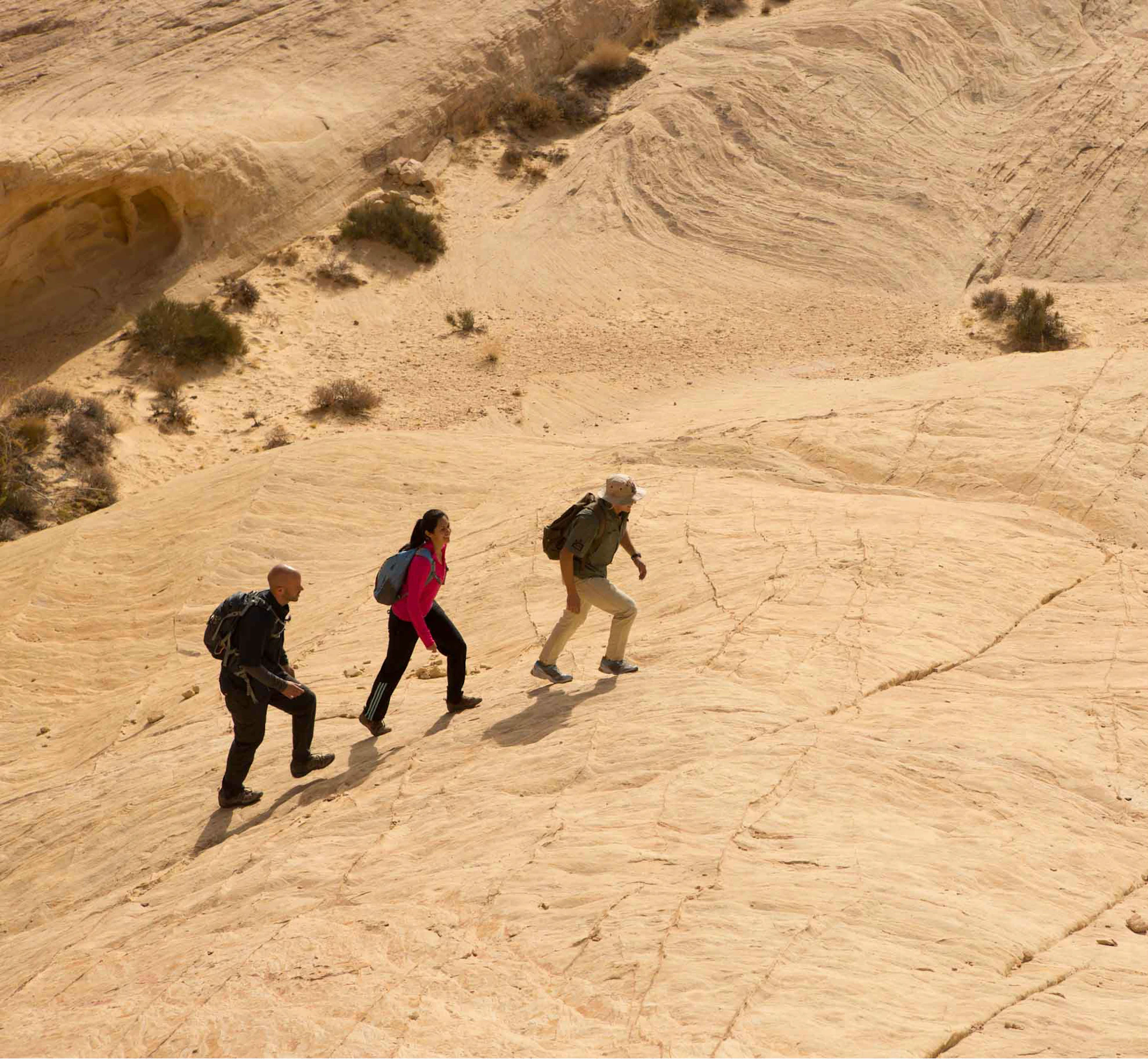 Three people with backpacks hike up a sandy, rocky slope in a desert landscape with sparse vegetation.