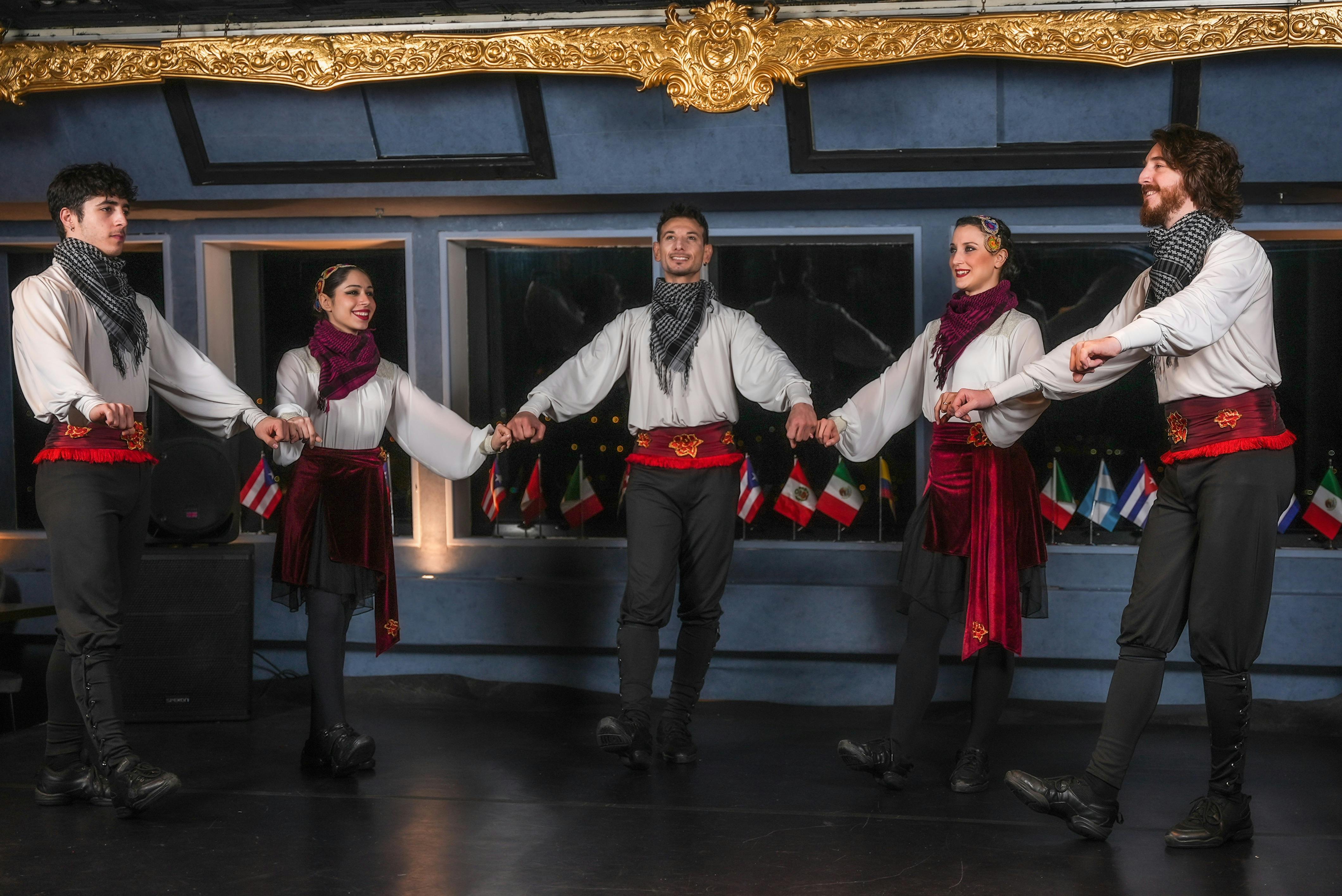 伝統的なダンスウェアを着た4人が手をつないで笑顔を見せ、背景には複数の旗が表示されています。