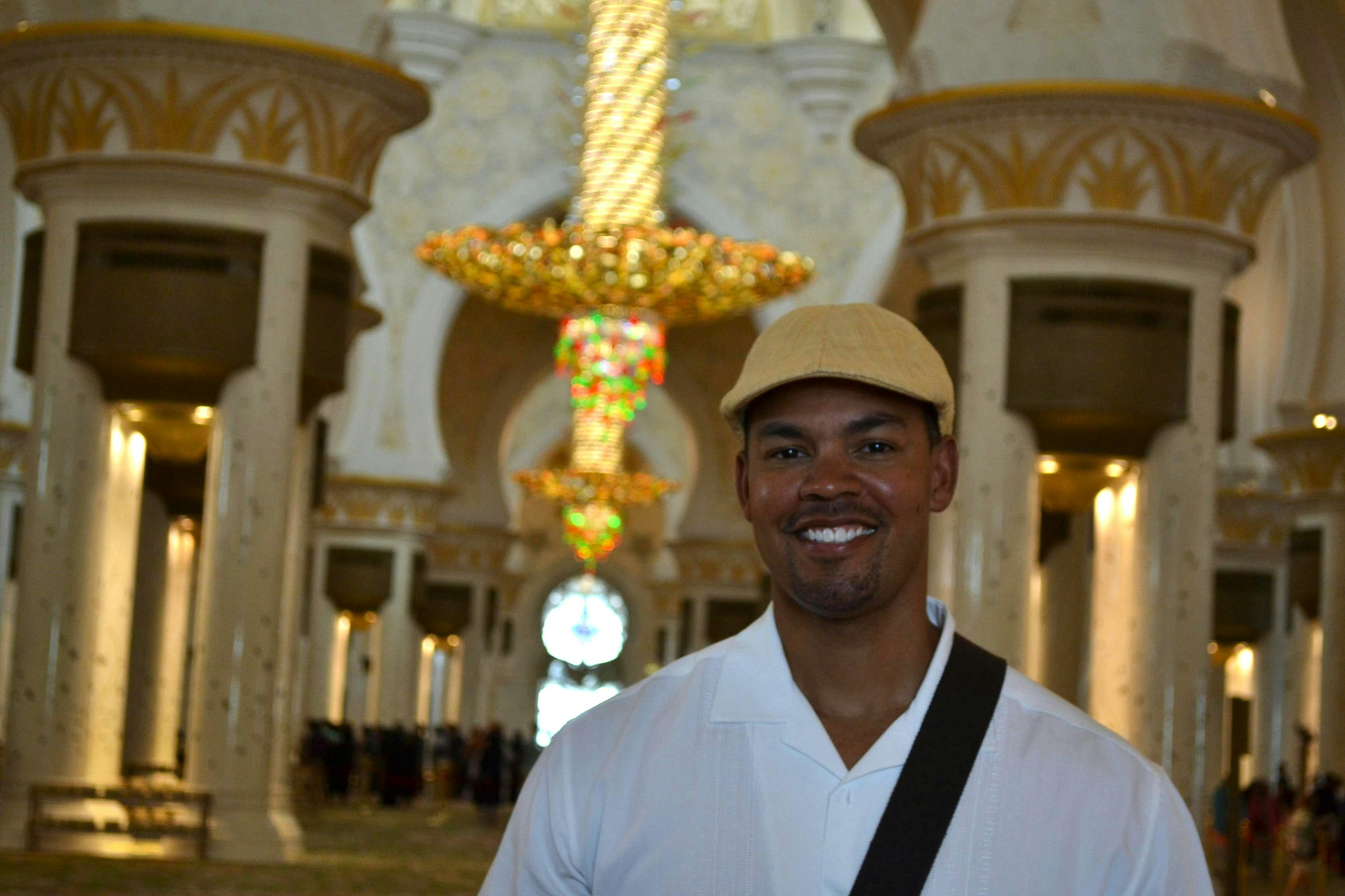 A person smiling in front of an ornate interior with a large chandelier and decorated columns. The person wears a beige hat.