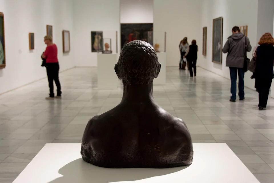A gallery with a bronze bust in the foreground and people viewing art on the walls and pedestals in the background.