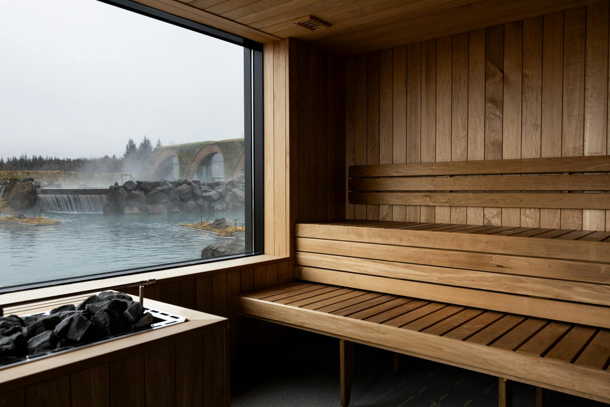 Wood-paneled sauna interior with a bench, large window overlooking a misty outdoor hot spring with rocks and an arched bridge.