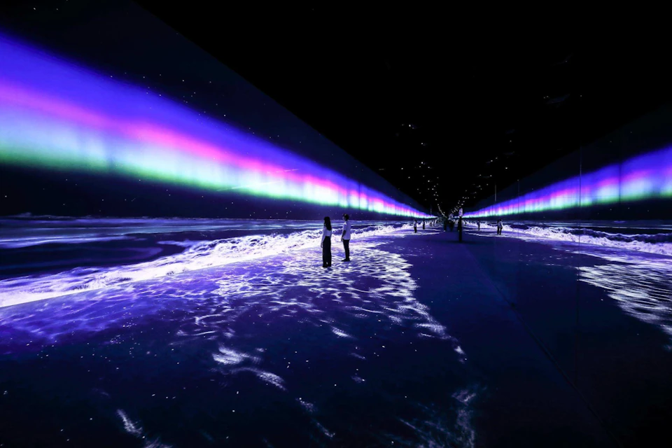 People standing on a reflective surface with projections of waves and an aurora-like light display in a dark space.