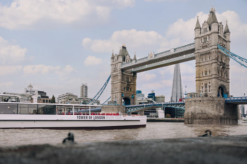 A view of Tower Bridge with a tour boat labeled 'Tower of London River Tour' on the Thames and the Shard in the background.