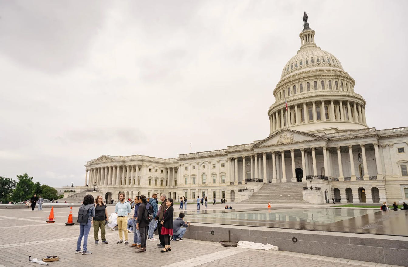 多云天，一群人站在美国国会大厦外面，柱子和穹顶清晰可见。