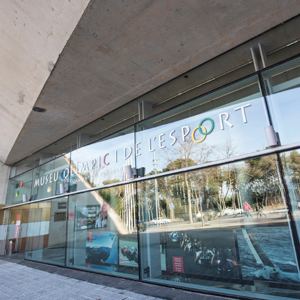 A modern building with large glass windows displaying signs for "Museu Olímpic de l'Esport" and reflections of trees outside.