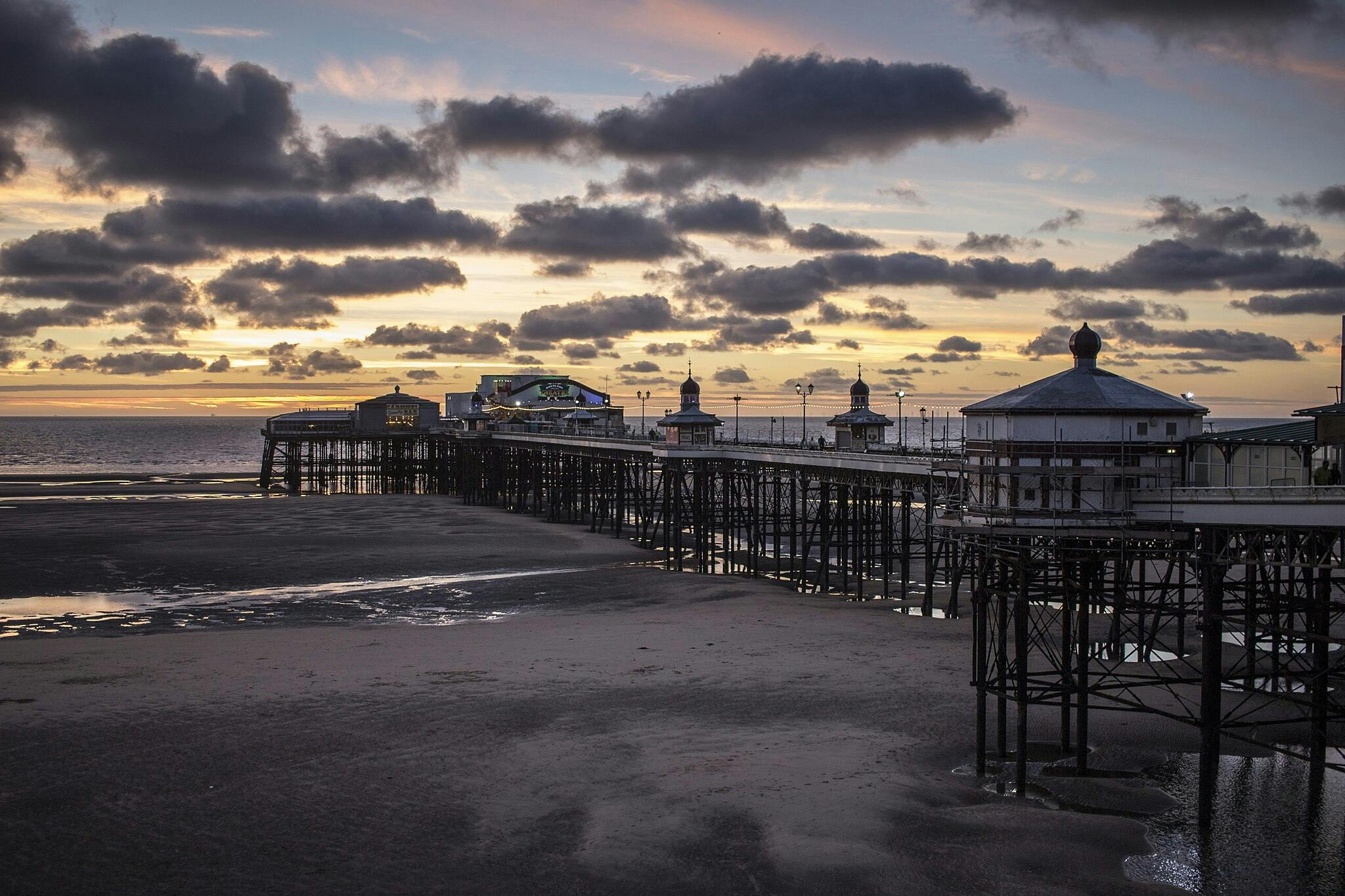Une jetée s'étend sur le sable mouillé jusqu'à l'océan sous un ciel nuageux de coucher de soleil, avec des bâtiments éclairés le long de sa structure.