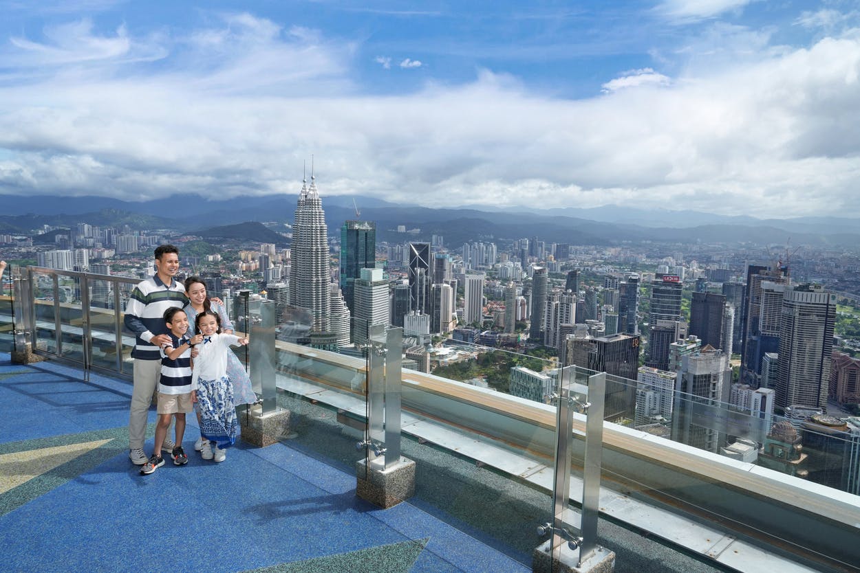 A family of four stands on an outdoor observation deck with a city skyline, including tall buildings, in the background.
