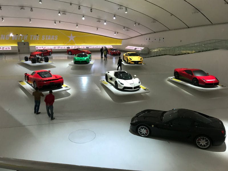 Une salle d'exposition présentant diverses voitures de sport, dont des modèles rouges, verts, blancs, jaunes et noirs, avec quelques personnes en train d'observer.