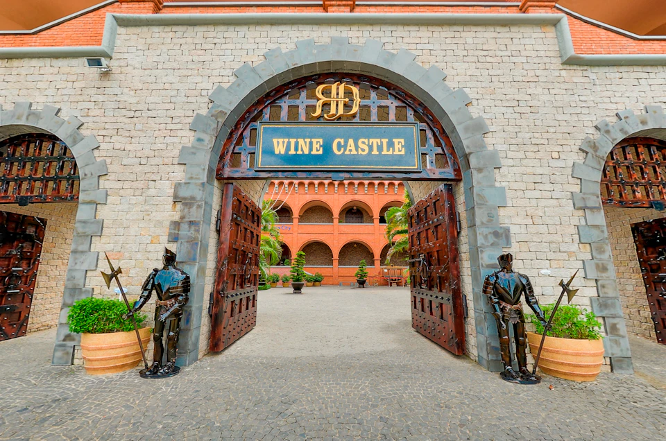 A stone archway with a sign reading 'Wine Castle,' flanked by knight statues, leading to a courtyard with red brick architecture.
