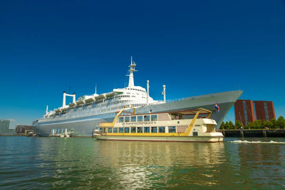 A large cruise ship is docked in calm waters with a smaller boat named "De Pannenkoekenboot" passing by in the foreground.