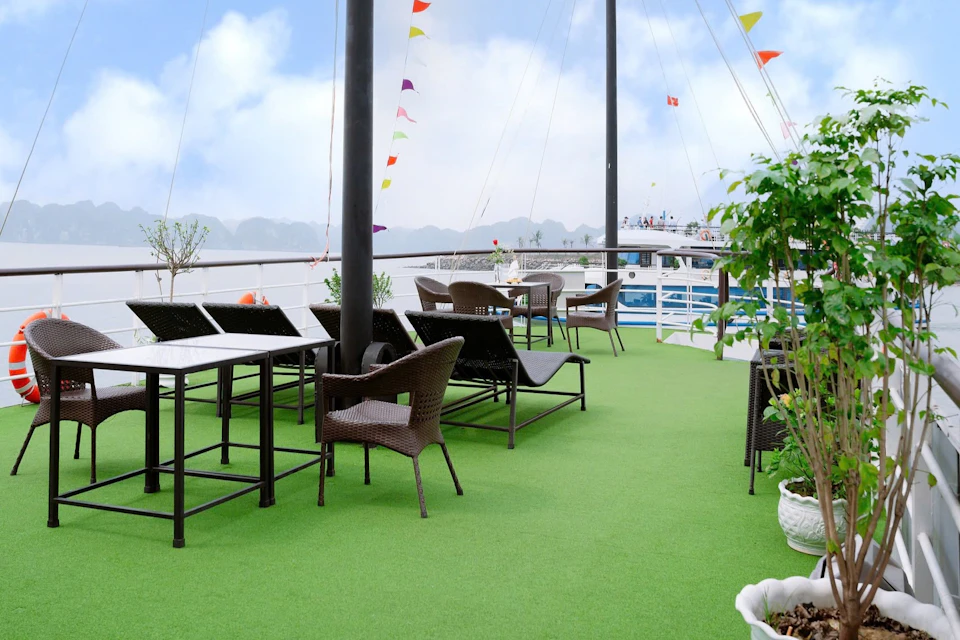 Outdoor deck with green flooring featuring wicker chairs, tables, potted plants, and colorful flags. Boat and mountains in the background.