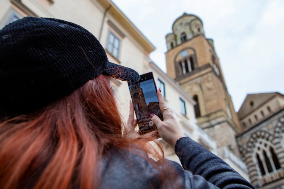 A person with red hair and a black cap takes a photo of a historic building with a bell tower using a smartphone.