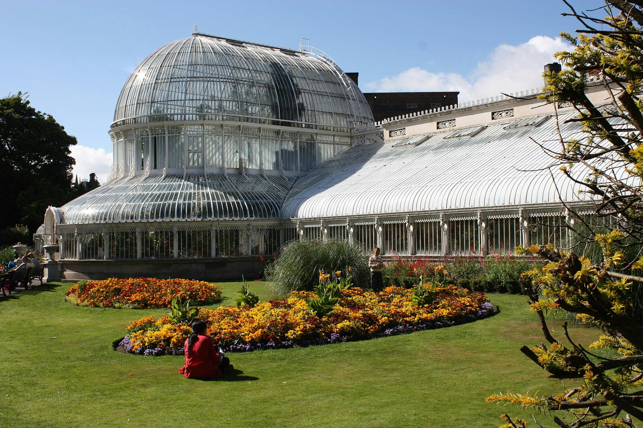 Un grand jardin d'hiver en verre avec des parterres de fleurs colorés devant, une personne en rouge assise sur l'herbe, et un ciel bleu clair au-dessus.