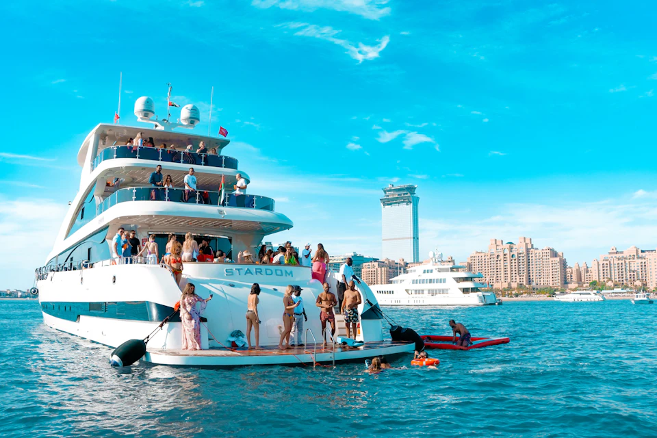 A group of people enjoying a sunny day on a large yacht named "STARDOM" with city buildings in the background.