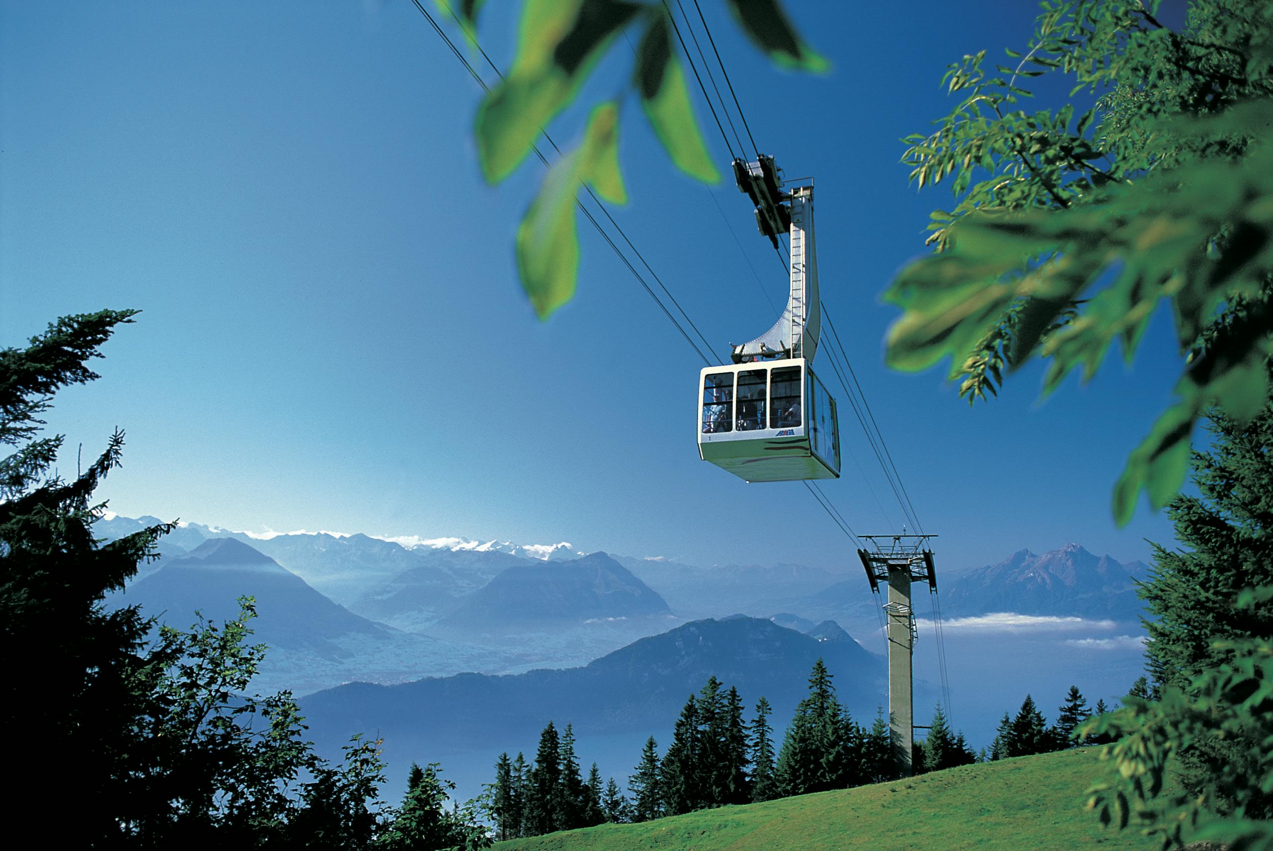 Nad zeleným svahem s horami a jasnou oblohou v pozadí projíždí lanovka.