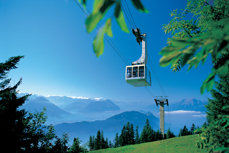 A cable car travels above a green hillside with mountains and a clear sky in the background.