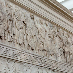 En detaljerad marmorfris som visar en procession av mantlade figurer huggna in i en vägg, med invecklade geometriska och blommiga mönster nedanför.