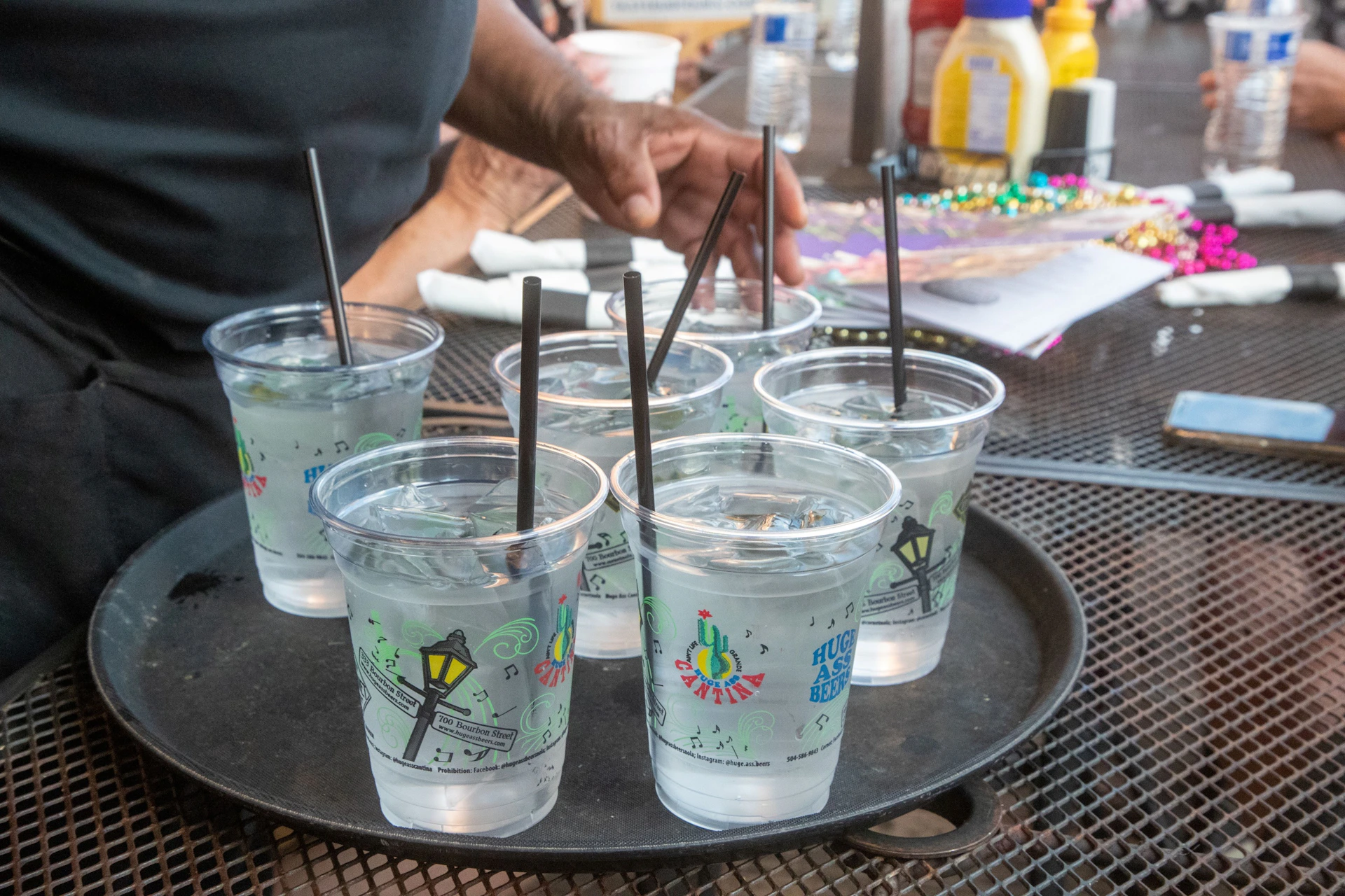 A tray holding six plastic cups of water with black straws, on a table with condiments, beads, and other items nearby.