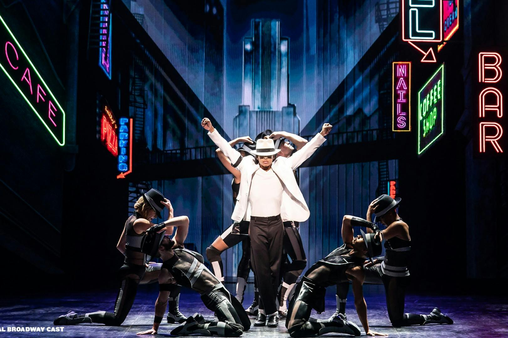 Um artista de fato branco e chapéu de feltro posa com bailarinos num palco iluminado a néon com sinais a dizer "CAFE", "BAR" e "COFFEE SHOP".