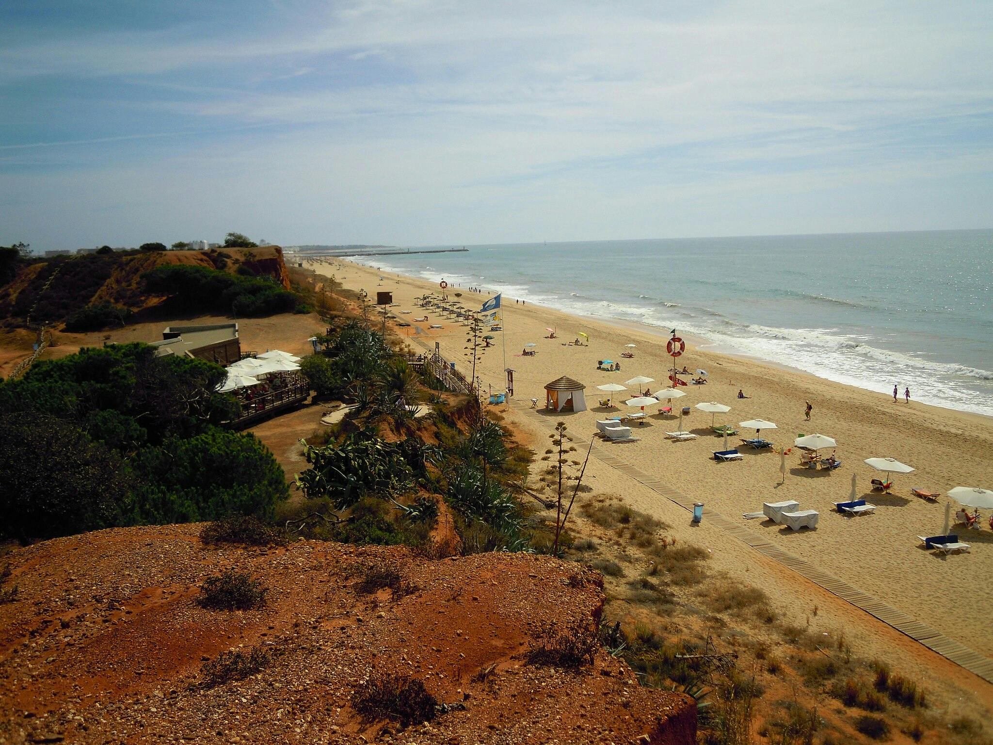 Uma praia de areia com guarda-sóis dispersos, espreguiçadeiras e algumas pessoas junto à costa. É visível uma falésia e edifícios junto à costa.