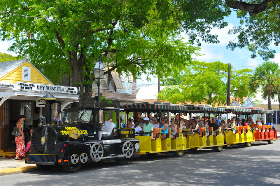 People riding on a yellow, open-air tourist train passing by shops and trees in a sunny, tree-lined street.