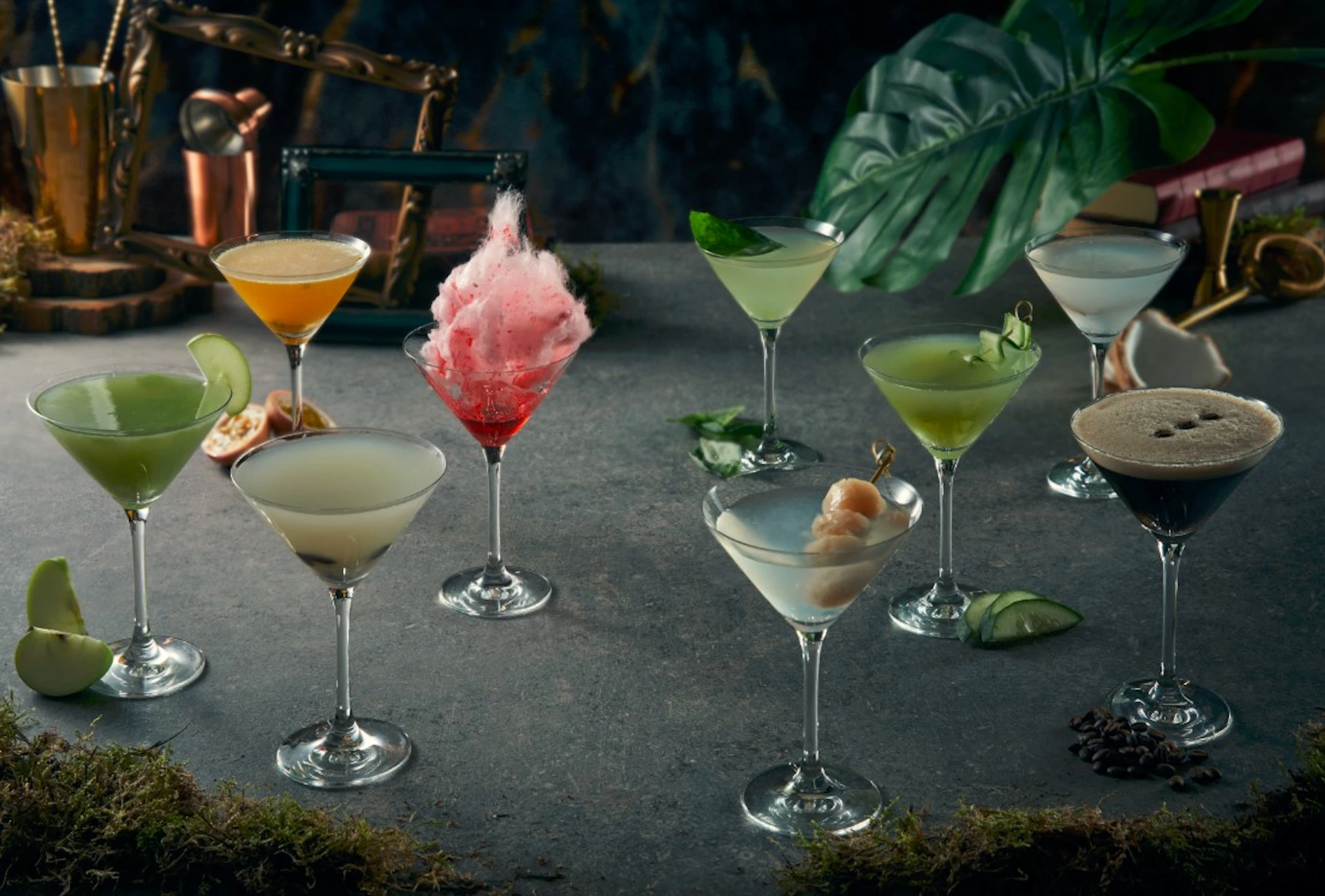 A variety of colorful cocktails in martini glasses on a gray surface with tropical leaves in the background.