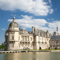 An ornate, historic chateau with turrets and intricate detailing sits beside a calm body of water under a partly cloudy sky.