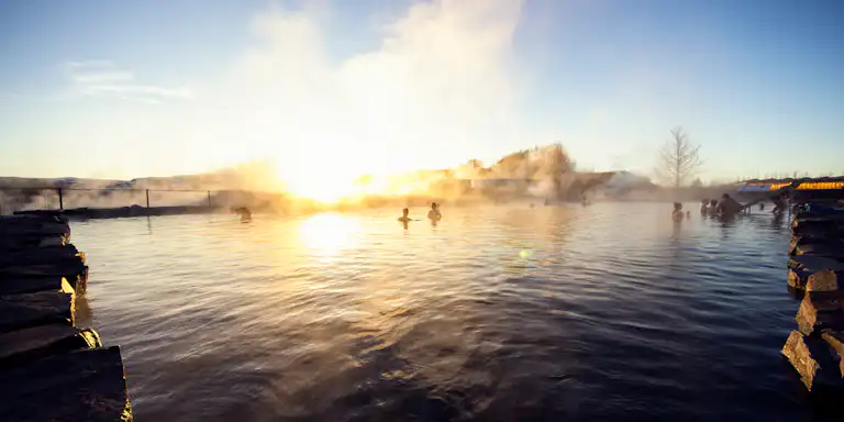 Menschen, die bei Sonnenuntergang in einer heißen Quelle stehen, umgeben von Dampf, mit Steinkanten und Bäumen im Hintergrund.