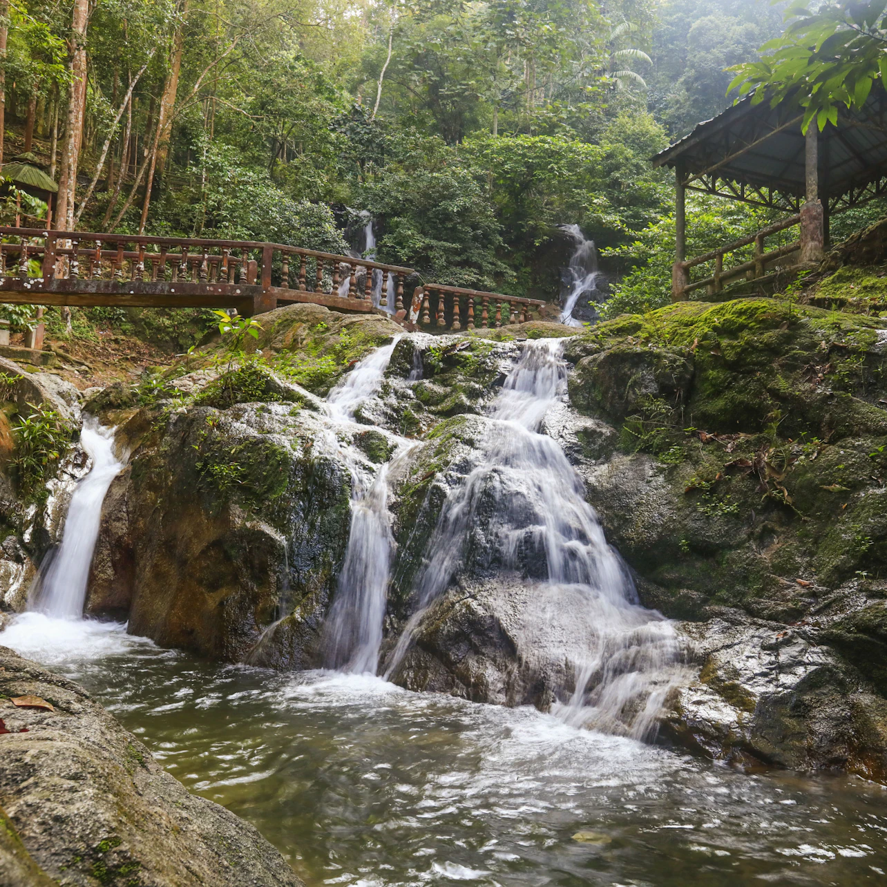 A small waterfall cascades down moss-covered rocks, surrounded by lush greenery. Wooden bridges and a gazebo are visible nearby.