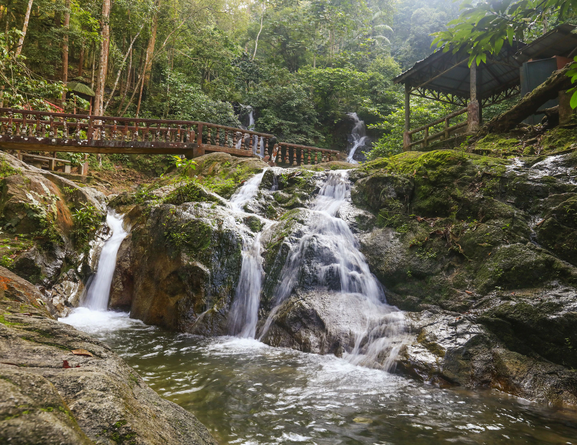 A small waterfall cascades down moss-covered rocks, surrounded by lush greenery. Wooden bridges and a gazebo are visible nearby.