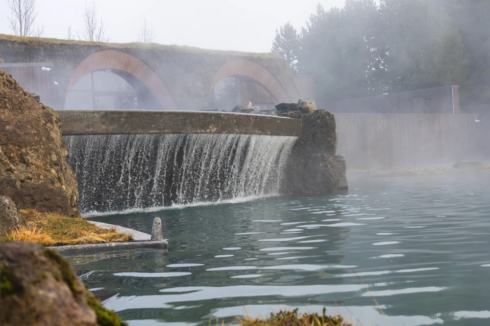 A small waterfall flows into a serene blue pool, with steam rising and arched bridges in the background under a misty sky.