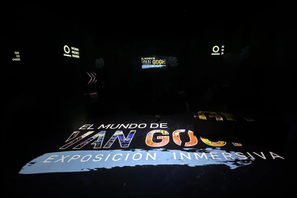 Dark room with illuminated text on the floor reading 'El Mundo de Van Gogh, Exposición Inmersa' and other signs visible.