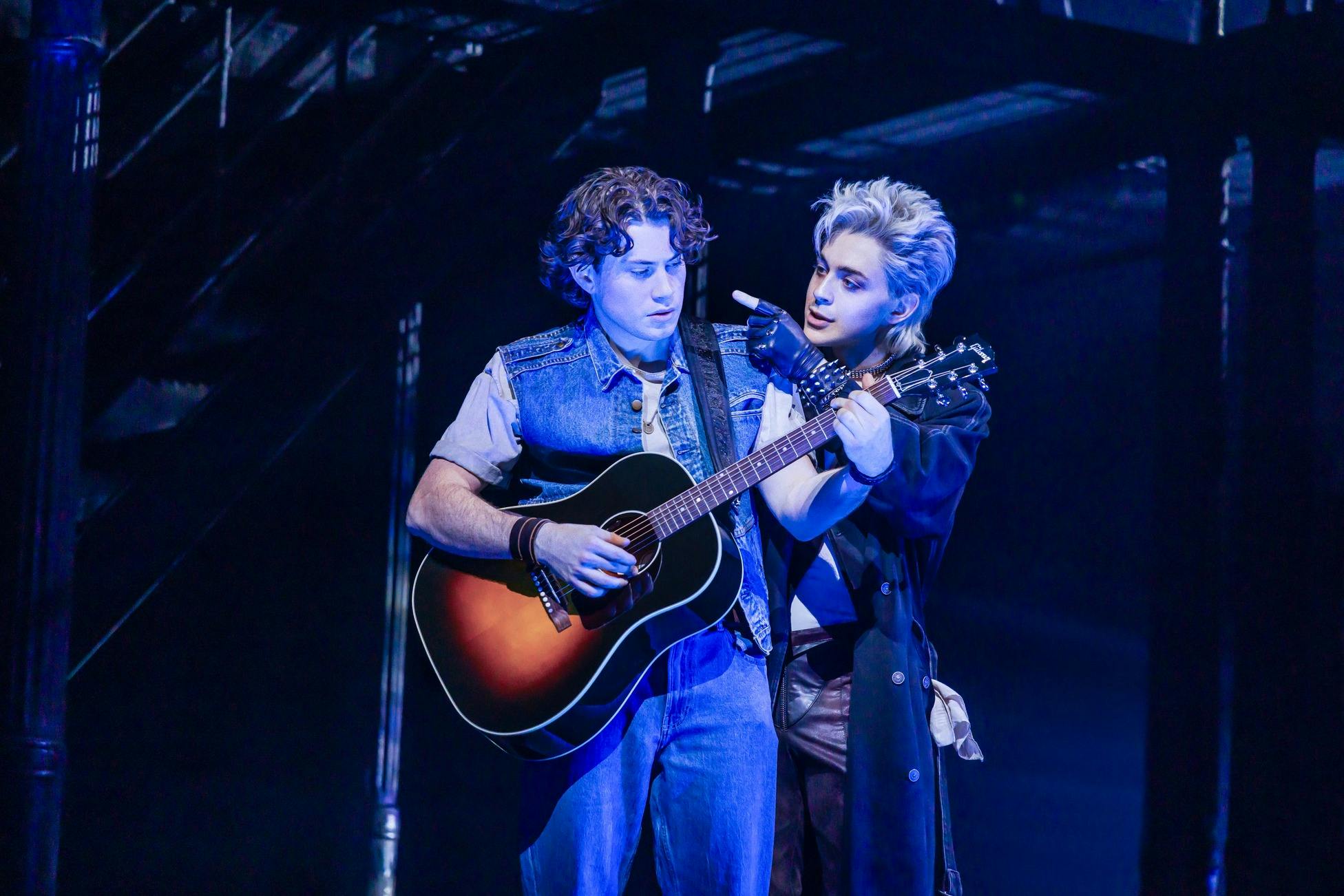 Two performers on stage, one holding a guitar, the other gesturing towards them, under blue lighting.