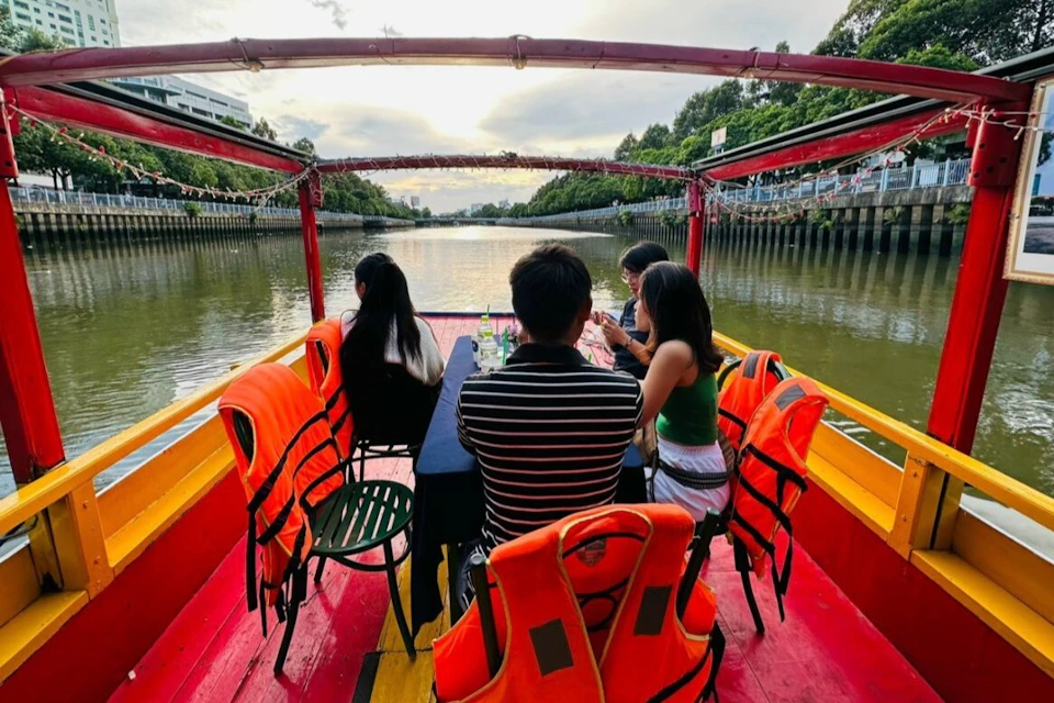 Four people sit on a boat with life jackets placed on chairs, cruising along a river lined with trees and buildings.