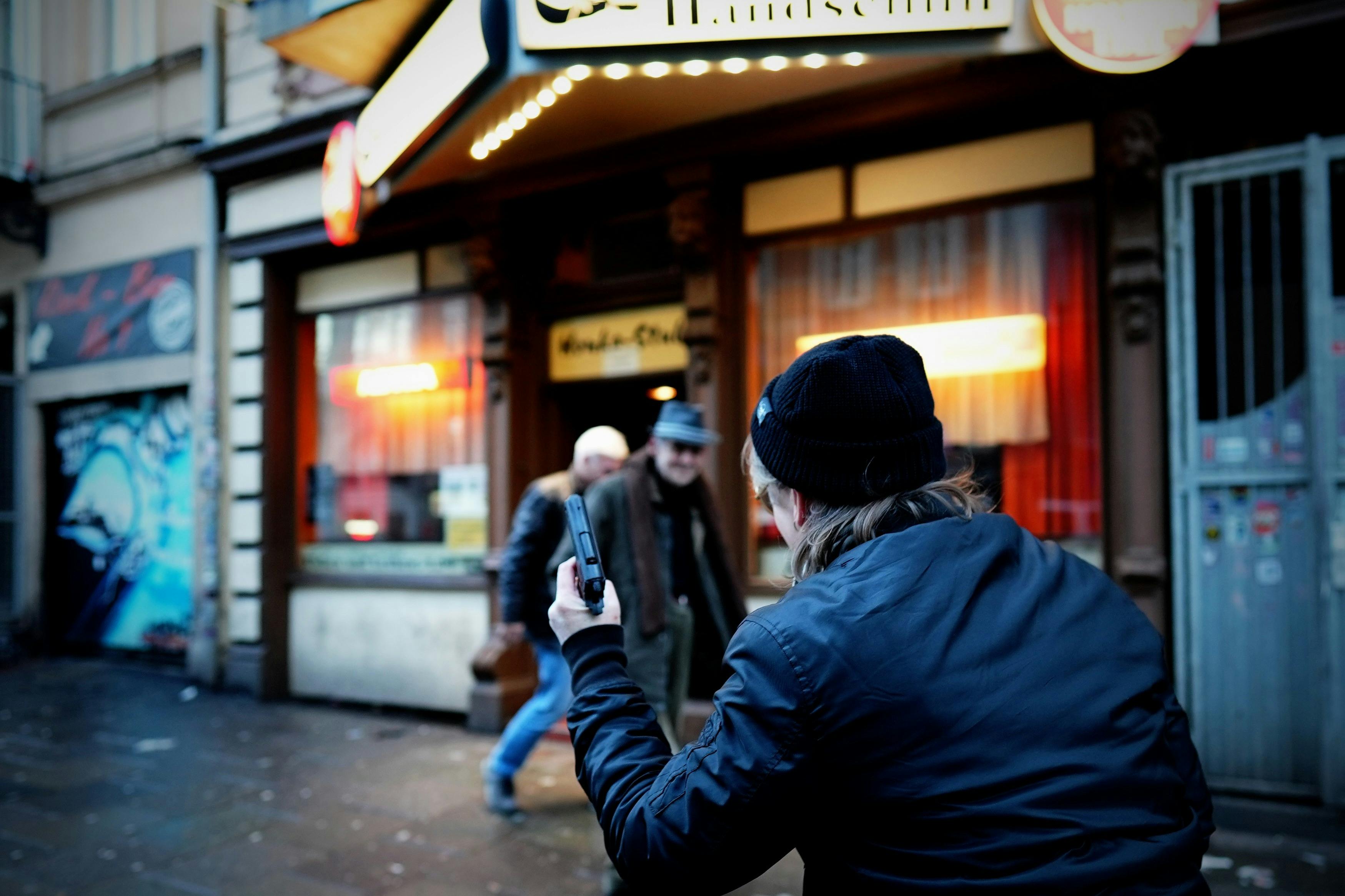 一个穿着便帽的人在一个灯光明亮的店面前拍摄两名男子，店面上铺着红色的窗帘和标志。