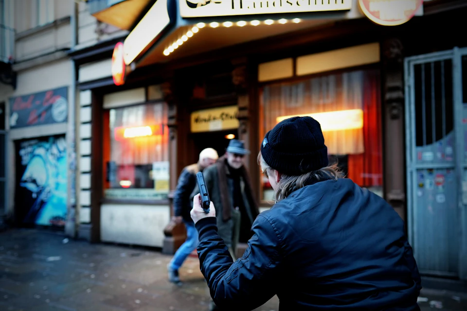 Person in a beanie photographing two men in front of a brightly lit storefront with red drapes and signs.