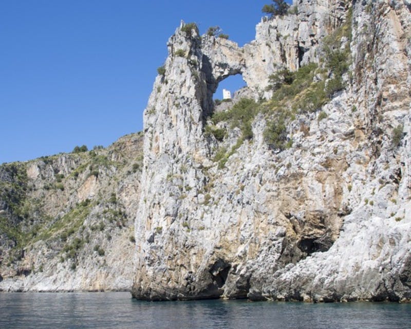 Steep rocky cliff with a natural arch formation and sparse vegetation, bordered by the sea under a clear blue sky.