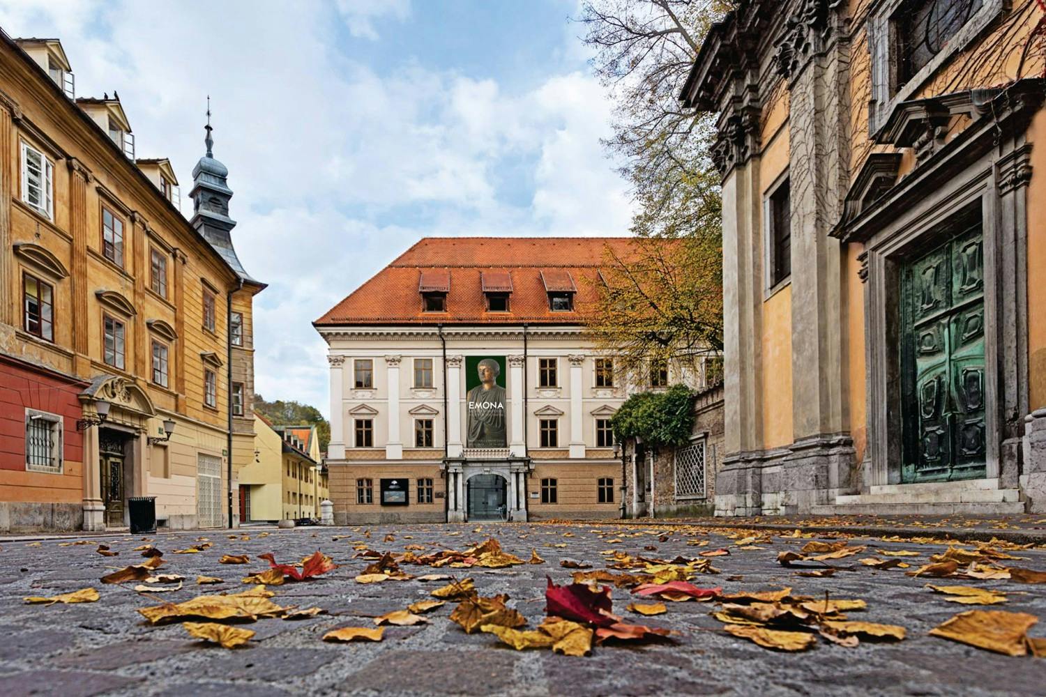 Dlážděné náměstí s podzimními listy, obklopené historickými budovami, včetně jedné s portrétním praporem na fasádě.