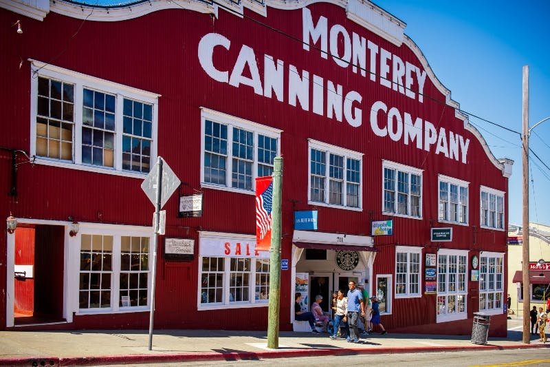 Red building with "Monterey Canning Company" signage, large windows, and an American flag. People walking on the sidewalk below.