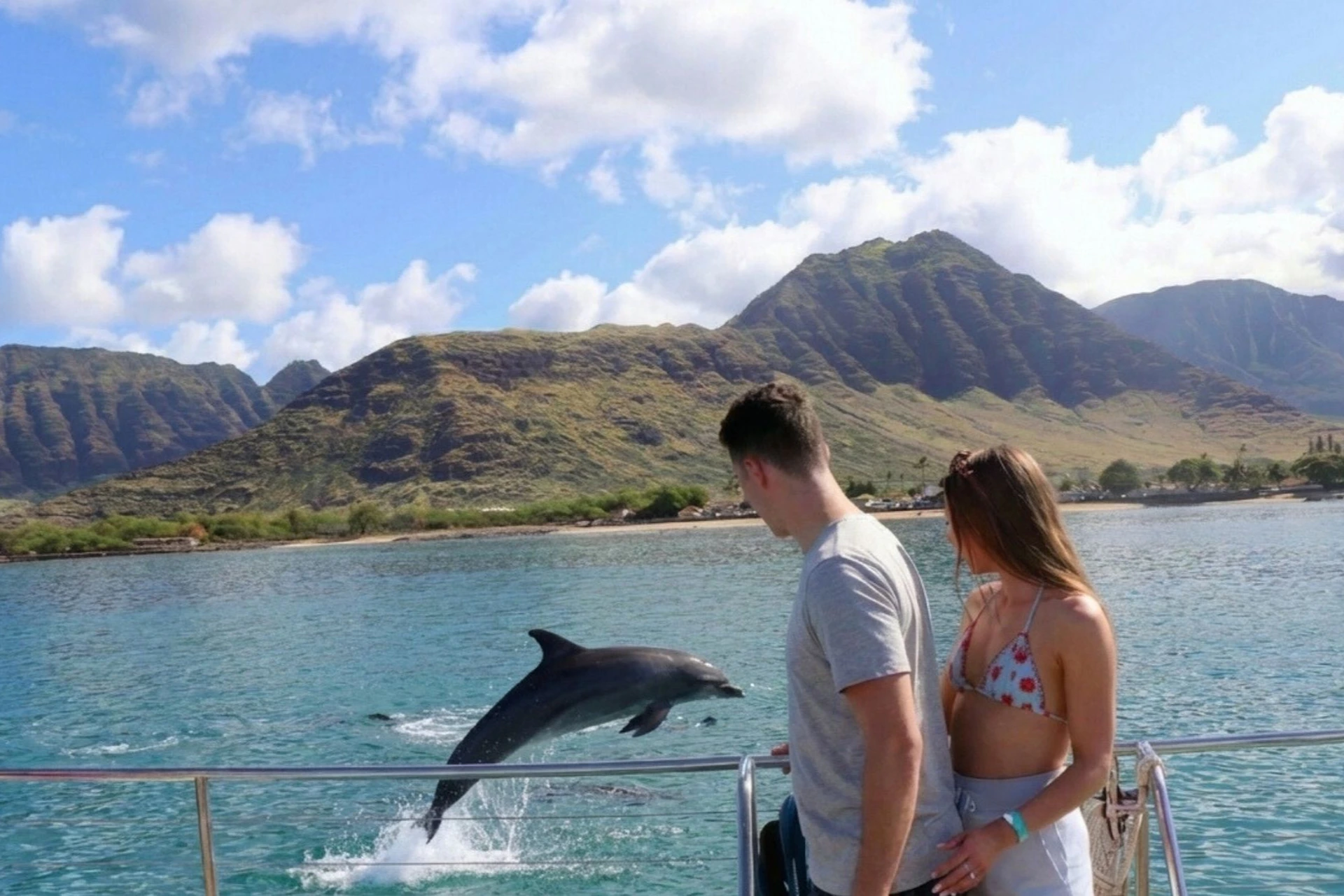 Two people on a boat observe a dolphin jumping out of the water near a scenic coastline with hills under a partly cloudy sky.